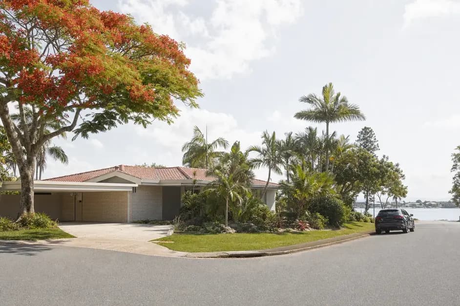 A single storey brick and tile home on a riverside street with wide eaves, a carport and mature poinciana and palms in the yard