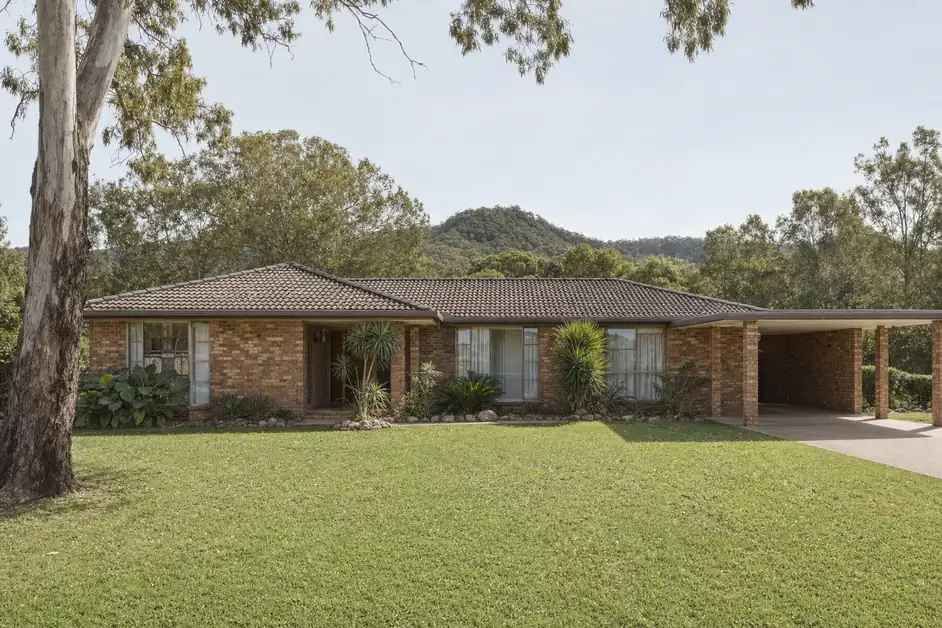 A 1980s low-set face brick house with a tiled hip roof on a large block, a carport to one side and forested hills in the distance