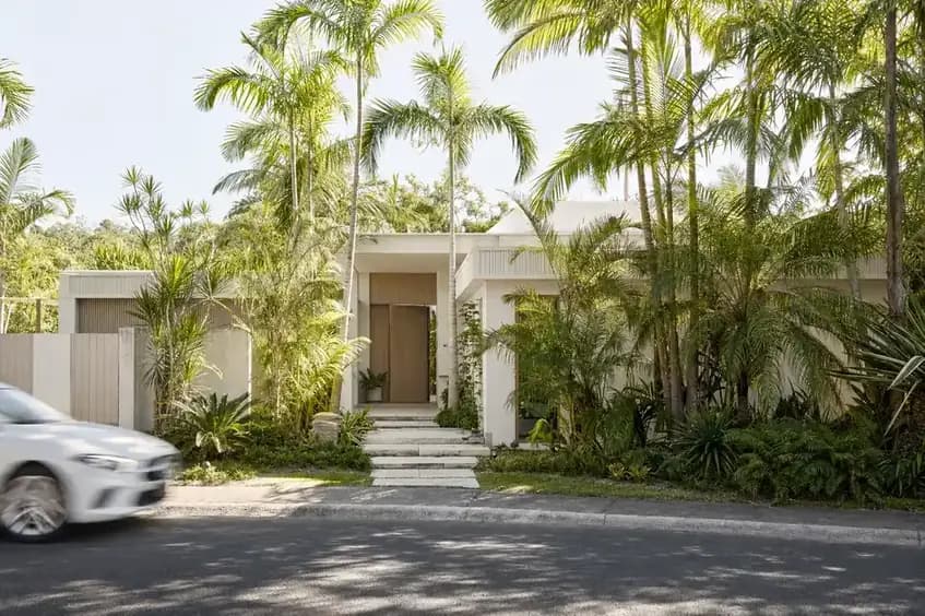 A low set contemporary home on a leafy Noosa street with white render, pale timber, deep roof overhangs and dense tropical planting screening the front
